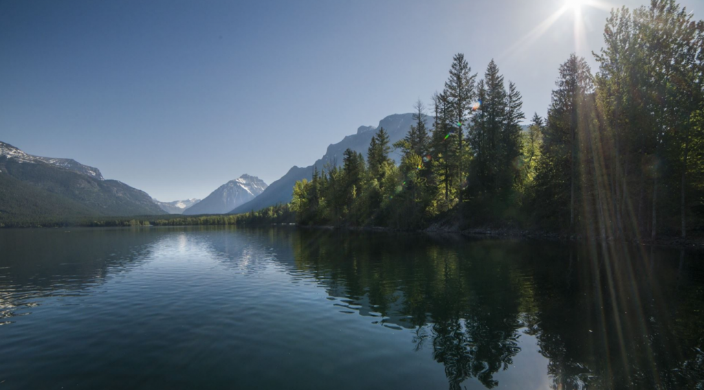 Image of a lake with mountains and trees in the background. The sun glints in the corner and the mountains and trees are reflected in the lake