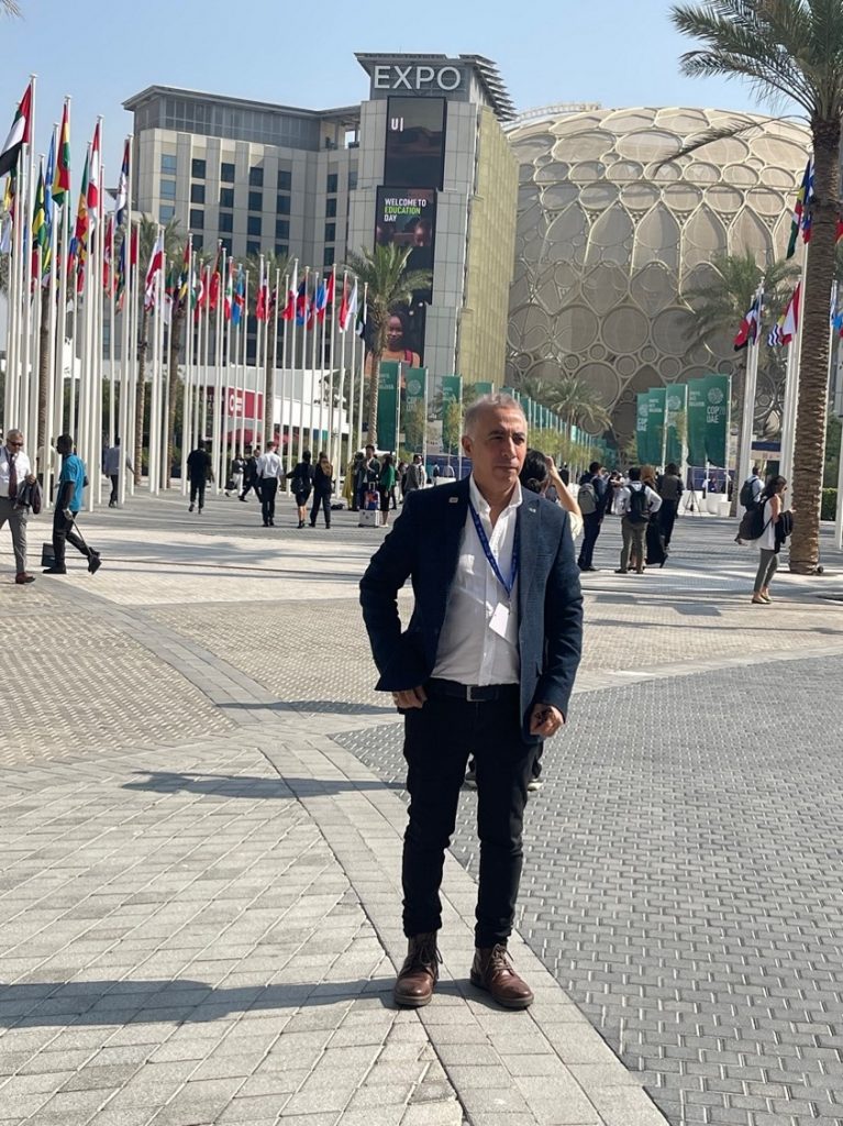 A man standing outdoors in a modern urban environment near a pathway lined with flags from various countries.