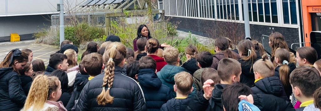 A group of children gathered outdoors around a woman speaking to them.