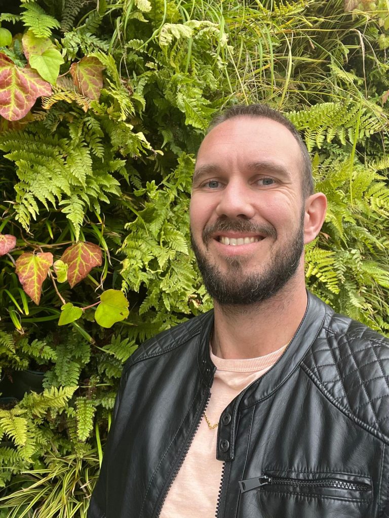 A man standing in front of a green wall covered with ferns and other plants.