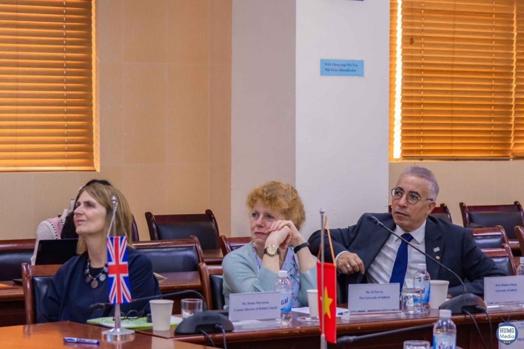 The image shows three people seated at a table during a formal meeting. Two flags, one representing the UK and the other representing Vietnam, are placed on the table.