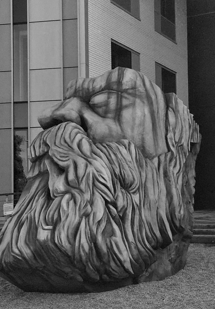 large grey statue of a fierce looking man with a large beard and top part of his head sliced off. it sits on a gravel floor in front of grey building.