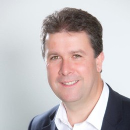 A headshot of Mark Bew. Middle aged white man with dark hair, smiling. He is wearing a white shirt with navy blue/ black suit.